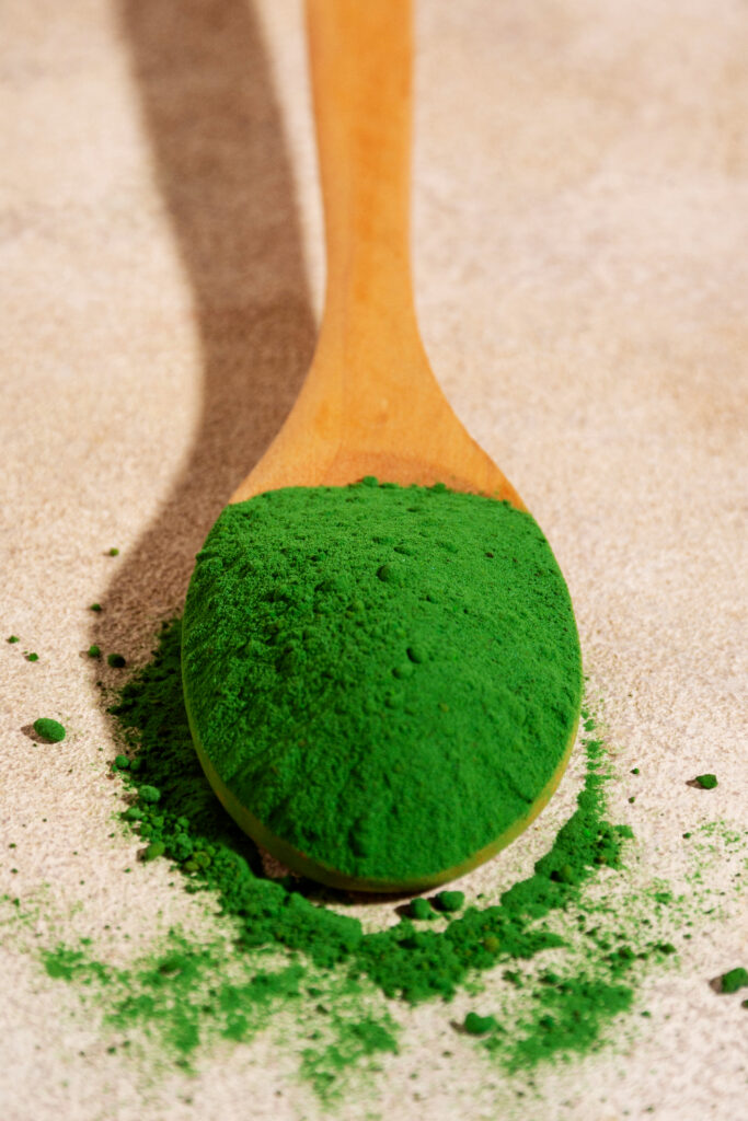 A wooden spoon filled with vibrant green greens powder, spilling slightly onto a beige textured surface.